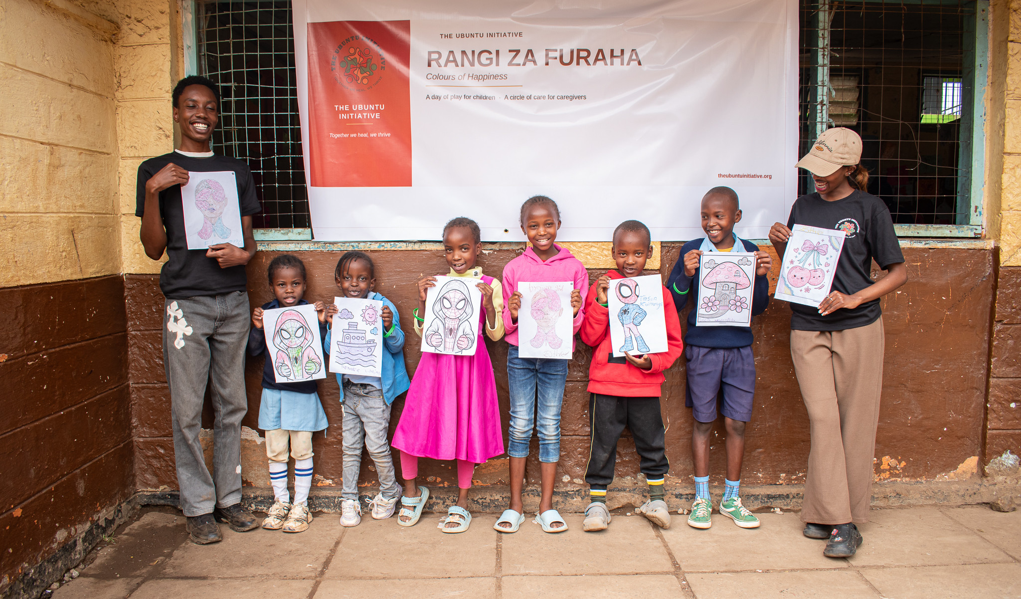 Children standing together outdoors at Kayole 1 Primary School, each holding up the artwork they made, with two Ubuntu volunteers beside them beneath the Rangi za Furaha banner